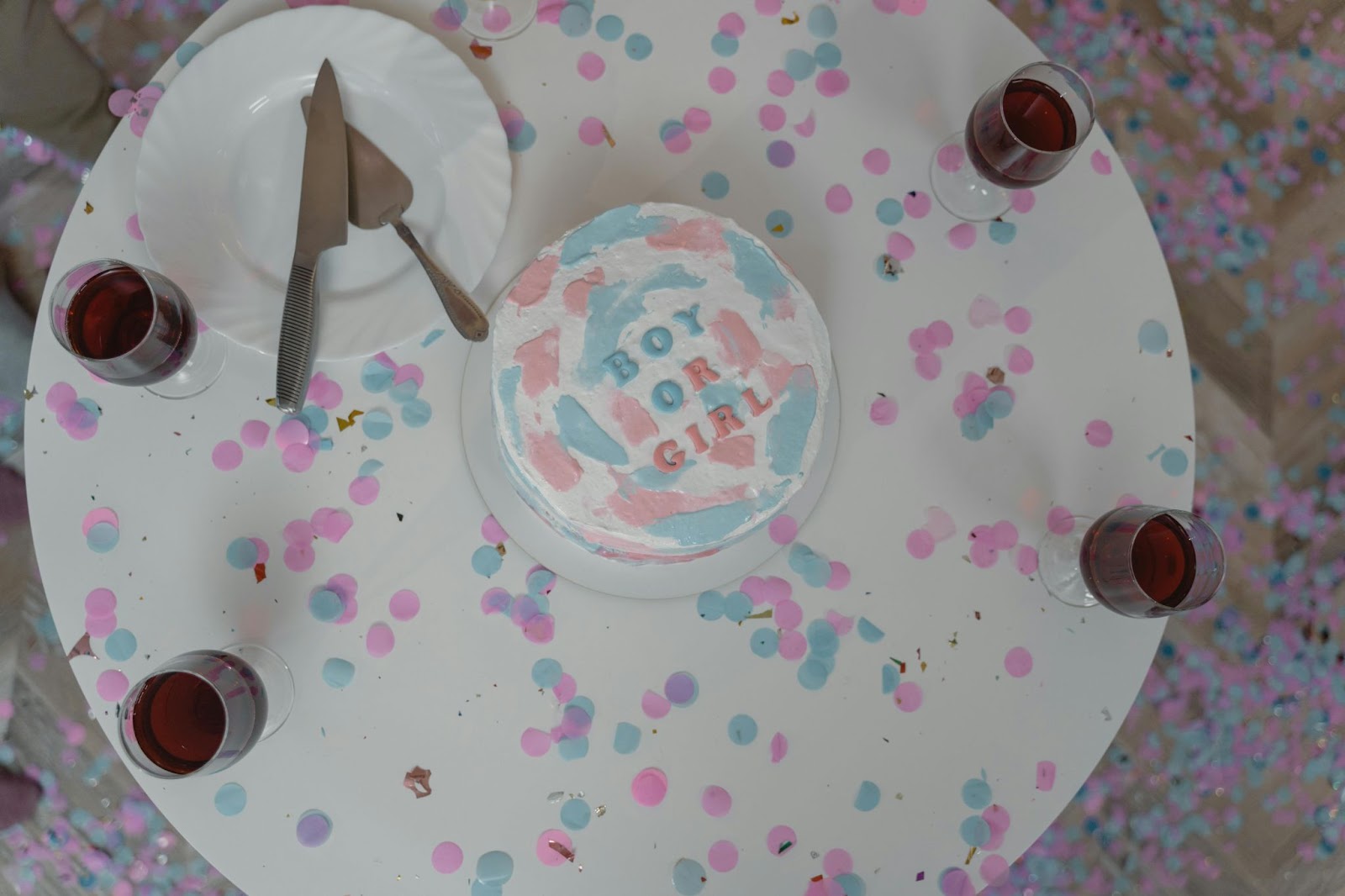 Top view of a gender reveal party cake with 'Boy or Girl' text, surrounded by pink and blue confetti on a white table.