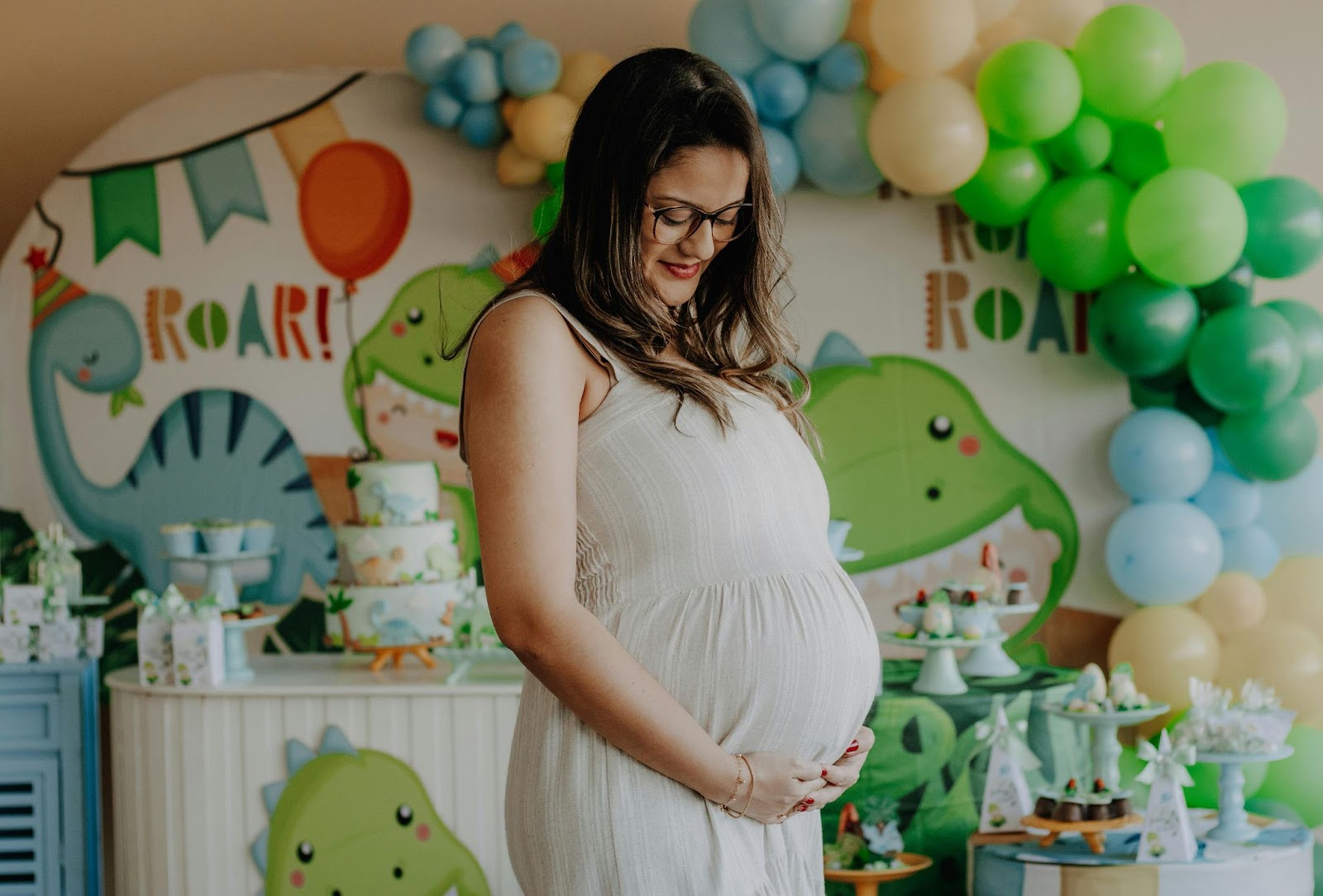 A pregnant woman in a white dress stands in front of dinosaur-themed decorations and balloons, showcasing planning a gender reveal party