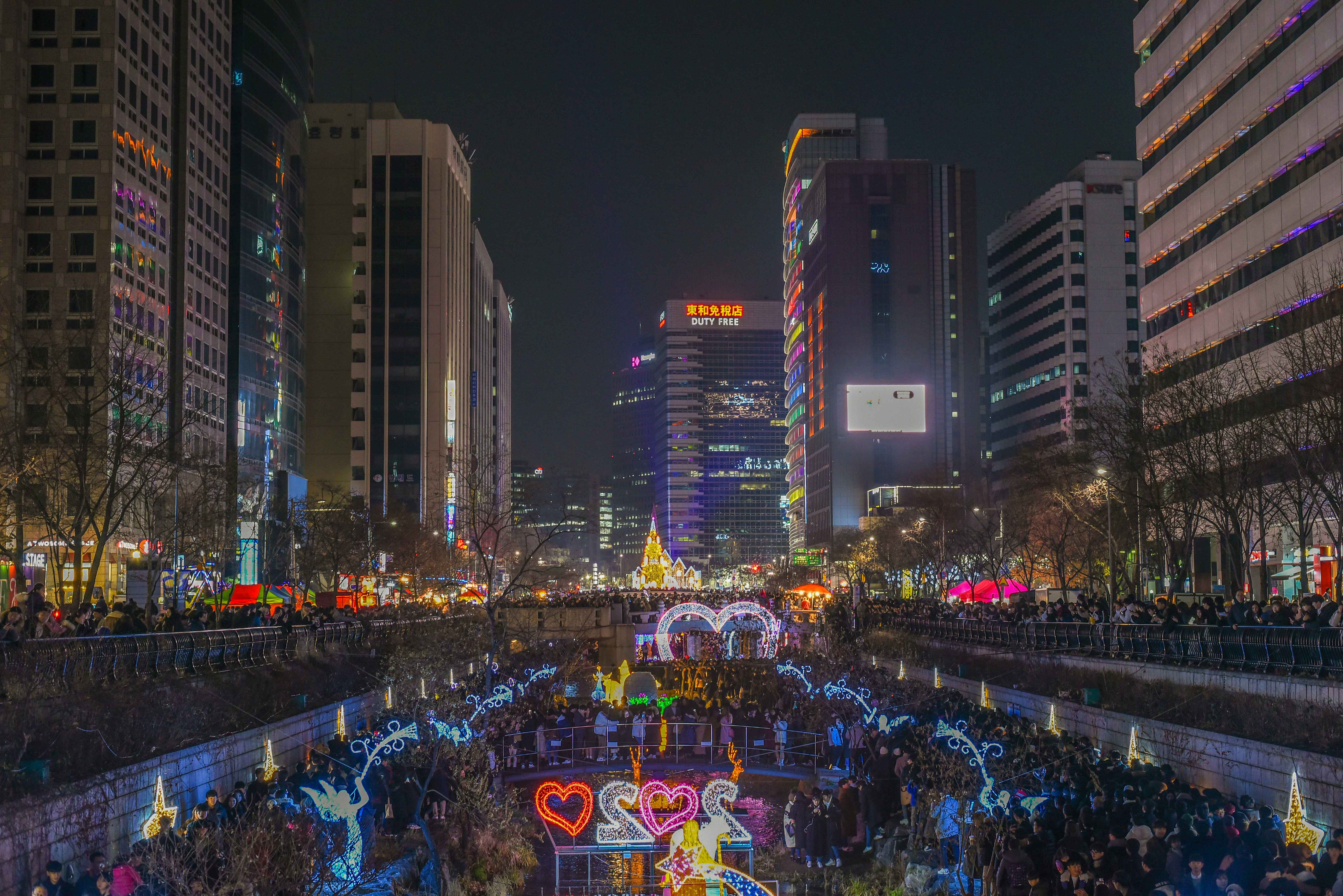 A festive nighttime scene with colorful lights, neon signs, and crowds near a canal decorated with heart-shaped and fairy-themed illuminations.